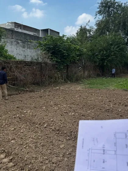 A site engineer holding the architectural plan while surveying the empty plot. This is where every project begins: translating the lines on paper to a vision on the ground.