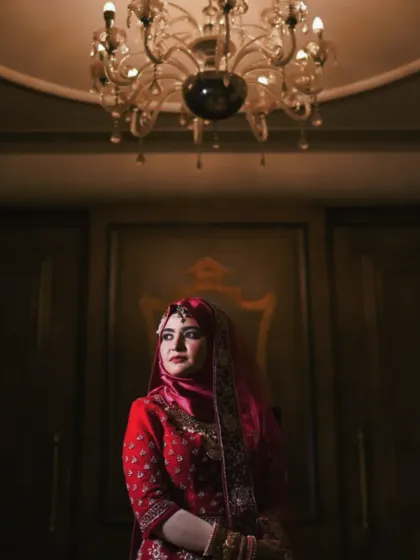 A portrait of a bride in her stunning red Nikkah attire. The dramatic lighting from the chandelier above creates a regal and elegant mood, highlighting her poise and traditional beauty.