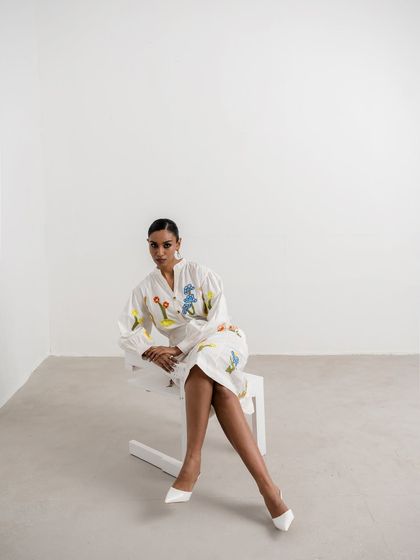 A seated portrait showcasing another embroidered white dress from the Nakateki collection. The styling with pointed white heels creates a sharp, polished, and sophisticated look.