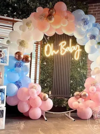A beautiful "Oh Baby" balloon arch over a faux greenery wall. The mix of pink, blue, and white balloons with rose gold accents is perfect for a sophisticated baby shower.