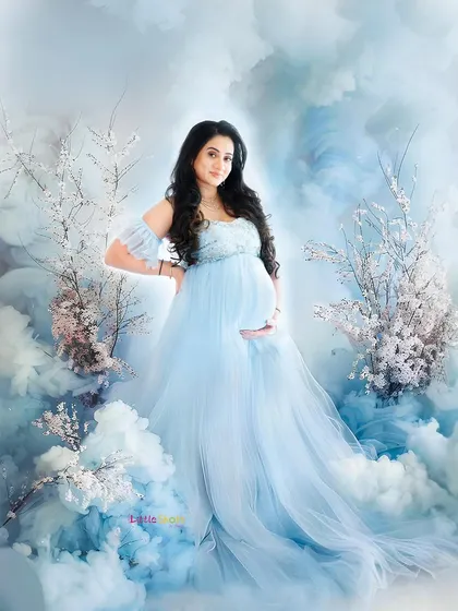 A full-length portrait from a dreamy, cloud-themed session. The mother-to-be, in a flowing blue gown, looks like a goddess standing among the clouds and delicate white branches.