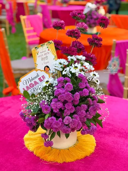 A vibrant centerpiece on a pink tablecloth, featuring purple and white flowers with a fun "Mehendi Laga Ke Rakhna" sign.
