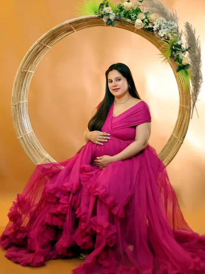 A mother-to-be in a vibrant pink ruffled gown, seated elegantly in a large circular prop decorated with pampas grass and flowers for a boho-chic studio look.