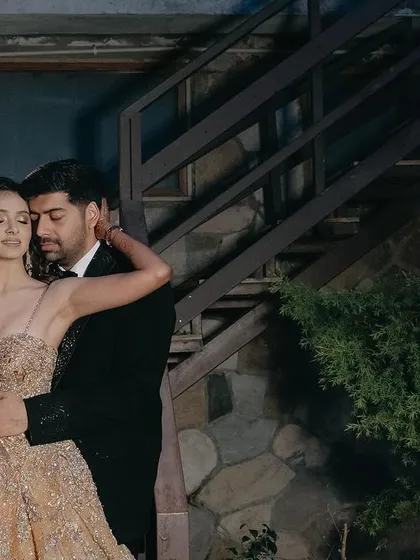 A glamorous outdoor portrait. The couple's embrace against the rustic stone and wood staircase creates a beautiful contrast, showcasing their elegant attire in a natural, romantic setting.