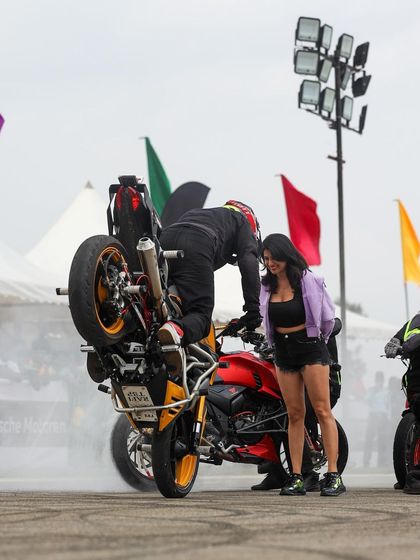 A stunt rider on a red bike leans into a trick, interacting with a model on the ground. Our stunt shows are choreographed performances that blend skill with showmanship.