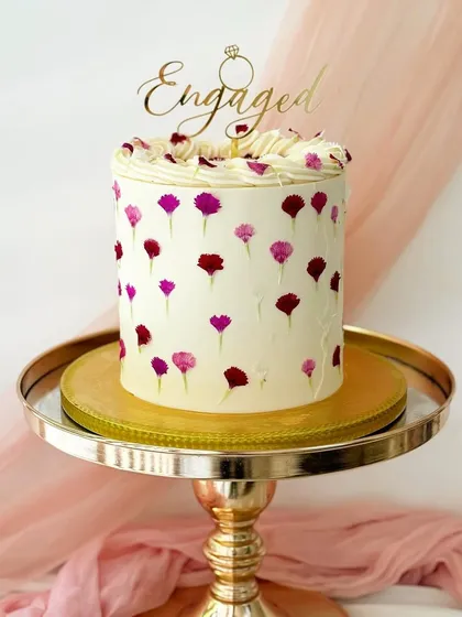 An elegant engagement cake featuring delicate, real pressed edible flowers pressed into smooth buttercream. A custom acrylic "Engaged" topper adds a personalized touch to this minimalist and natural design.