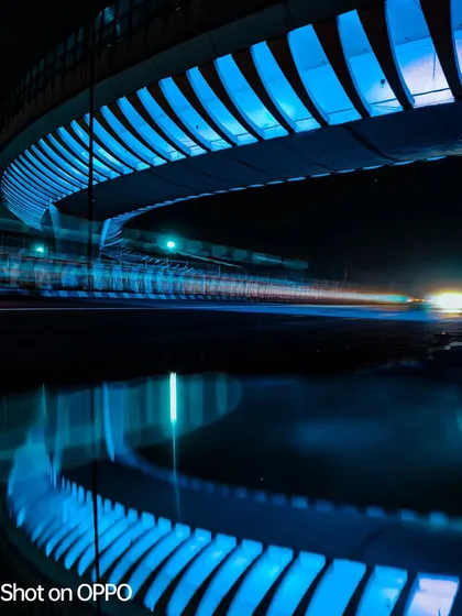 A long-exposure shot of a bridge at night, captured on an Oppo phone. The reflection in the water and the vibrant blue light create a serene yet futuristic urban scene.