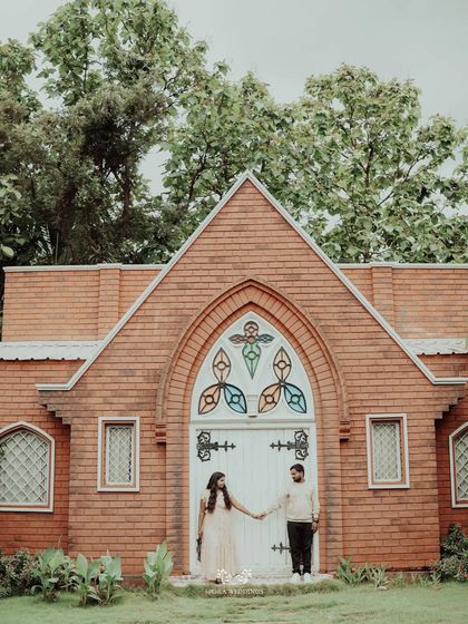 The couple holding hands in front of a beautiful brick building with stained-glass details.