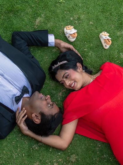 An artistic overhead shot of a couple lying on the grass. The mom-to-be is wearing a vibrant red gown with a cape, and the fabric spreads out beautifully around her.