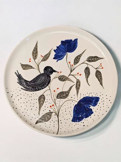 This sold plate showcases a bird nestled among leaves and bold blue flowers. Each piece is a unique, handcrafted creation, perfect for adding an artistic touch to modern homes.