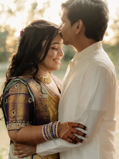 A tender and happy moment between a couple dressed in traditional South Indian attire. The groom kisses the bride's forehead as she smiles, bathed in warm, beautiful light.