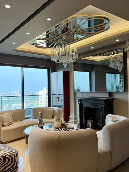 An overview of a luxurious living room in Gulshan Dynasty, Noida, where classic and modern elements converge. The space features plush, curved sofas, a statement marble fireplace, and a stunning mirrored ceiling detail that enhances the room's grandeur and reflects the city skyline.