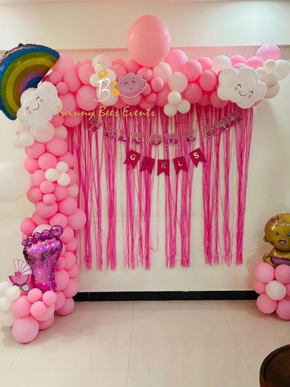 A fun and vibrant "Welcome Girls" decoration for a baby's homecoming. This all-pink theme uses a streamer backdrop, a full balloon arch with cute cloud and rainbow accents, and a baby girl foil balloon.