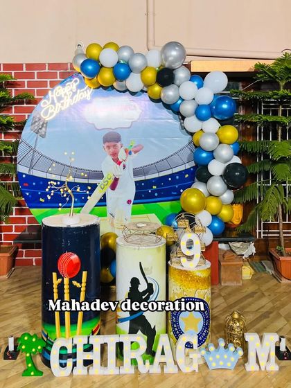 A creative cricket theme first birthday party for a young fan. This outdoor setup includes a custom backdrop showing a stadium, themed pedestals with cricket motifs, and a blue, yellow, and silver balloon garland.