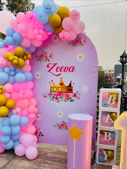 A lovely princess-themed first birthday for Zeeva on a terrace. The setup includes a personalized backdrop with a crown, a colorful balloon garland, 'BABY' blocks, and a light-up number '1'.