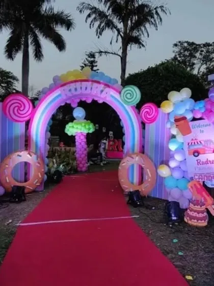 A spectacular 'Candyland' themed party entrance. This outdoor setup features a grand arch decorated with giant lollipops and donuts, a red carpet, and colorful lighting to welcome guests.