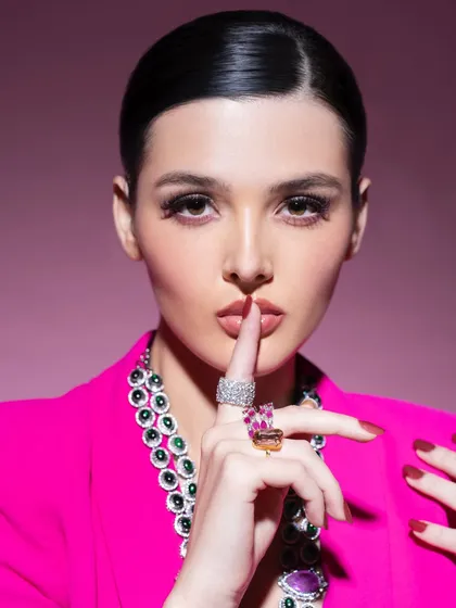 A powerful and intriguing shot from a high-fashion editorial. The styling combines a bold pink blazer with a statement multi-gemstone necklace and vibrant rings, creating a look that is both playful and commanding.