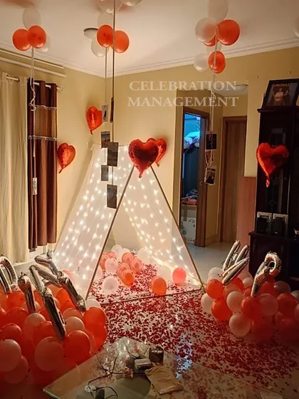 A cozy and personal surprise featuring a lit-up canopy tent in the living room. The floor is covered with rose petals, and photos of special moments hang from the ceiling balloons, making it a walk down memory lane.