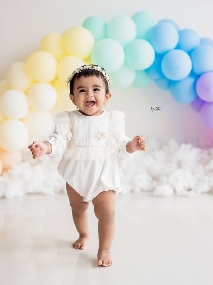 A joyful baby girl walking in front of a rainbow balloon arch for her first birthday shoot.