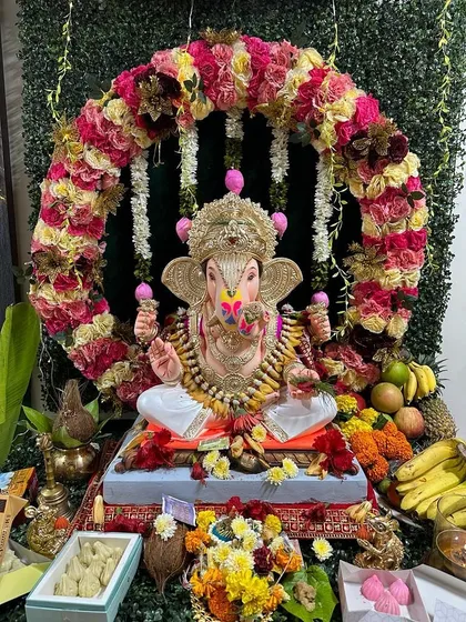 A circular floral arch made with multi-colored roses frames the Ganpati idol beautifully. The base is decorated with a variety of fruits and sweets, completing the traditional offering setup.