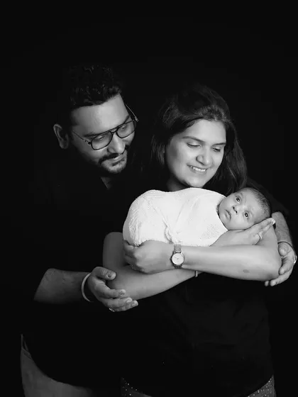 A dramatic black and white portrait of a new family. The low-key lighting creates an intimate and timeless feel, focusing entirely on the parents and their newborn.