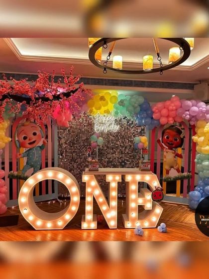 An elaborate Cocomelon-themed first birthday stage. This decoration features a rainbow balloon arch, a shimmer wall, cherry blossom tree props, and large "ONE" marquee letters.