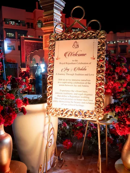 A welcome sign with ornate gold framing, introducing guests to the "Royal Rajputana Celebration." The sign is surrounded by deep red florals, setting a regal tone from the very beginning.