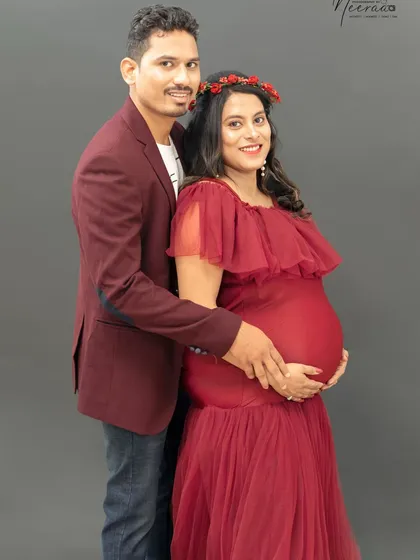 A beautiful, classic portrait of the couple. They stand together, hands lovingly placed on the baby bump, smiling warmly for the camera. The rich red gown is a perfect choice.
