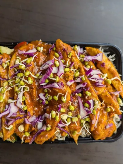 A close-up of a packed meal box, showing tender chicken strips in a savory sauce over a bed of rice, topped with fresh red cabbage and sprouts.