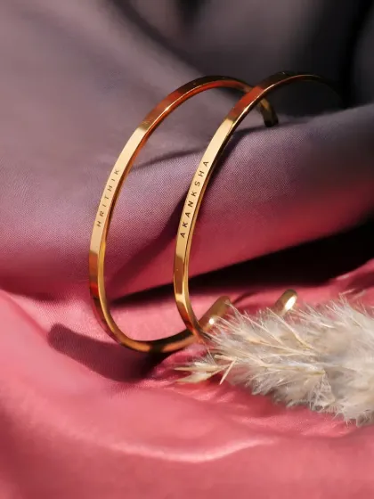 Two gold-plated cuff bracelets, engraved with 'HRITHIK' and 'AKANKSHA', displayed on soft fabric. This set is ready to be gifted to a special couple.