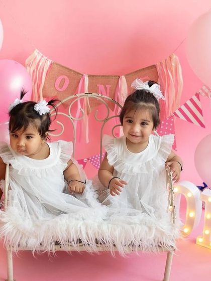 Twinning and winning! These adorable twin sisters are all smiles during their first birthday celebration in the studio, surrounded by pink and white balloons.
