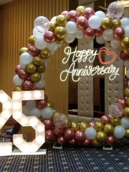Celebrating a 25th anniversary with style. This decor includes a circular balloon arch in rose gold, gold, and white, a 'Happy Anniversary' neon sign, and large marquee numbers, perfect for creating a memorable photo opportunity in a party hall.