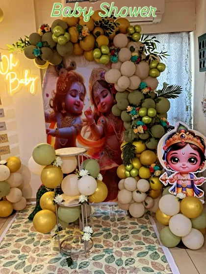 A unique Krishna-themed baby shower. The decoration features a backdrop with cute illustrations of Krishna and Radha, surrounded by a green and gold balloon arch.