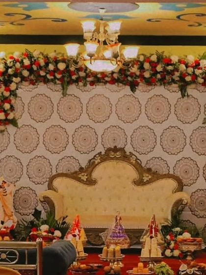 A traditional stage setup combining a patterned fabric backdrop with a heavy floral garland of red and white roses. The decor includes cow figurines, brass lamps, and flower vases, elements often used in auspicious ceremonies like housewarmings or baby showers.