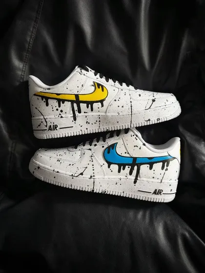 A striking pair of custom Nike Air Force 1s with a dual-color design, featuring a yellow dripping swoosh on one shoe and a blue one on the other, set against a black splatter background.
