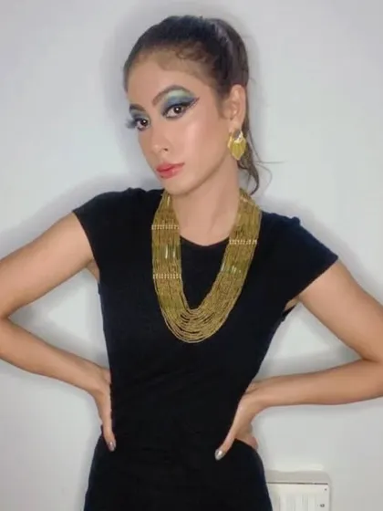 A full-length shot of a glamour fashion look. The makeup features a dramatic blue and green winged eyeshadow, complemented by a bold gold necklace and a sleek high ponytail for a modern, Cleopatra-inspired vibe.