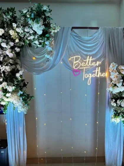 An elegant 'Better Together' backdrop perfect for an engagement or anniversary party. We use white drapes, fairy lights, a neon sign, and lush white artificial flower arrangements.