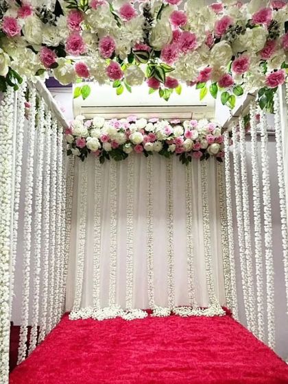 A four-poster mandap design with a canopy of pink and white flowers. The posts are draped with strings of white flowers, creating a beautiful and full look over a red velvet base.