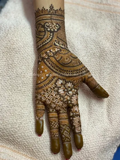 A close-up of a hand with a dense, intricate mehendi design. The pattern features lotus motifs and detailed grid work, showcasing a high level of skill.