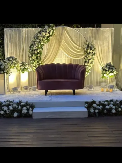 A chic and modern stage for a ring ceremony or reception. The design features a semi-circular floral arch, elegant white drapes, and a stylish velvet sofa.