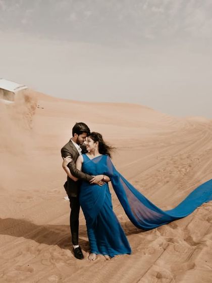 An epic, wide-angle shot in the desert, capturing the scale of the landscape and the intimacy of the couple. The flowing fabric adds movement and drama.