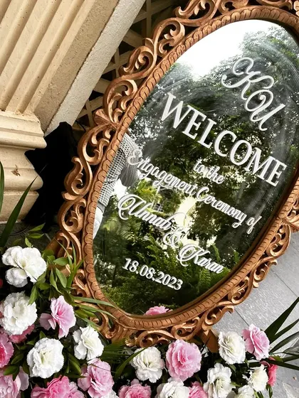 A unique and elegant welcome sign crafted on an ornate, vintage-style mirror for Unnati and Karan's engagement. The reflection of the surrounding greenery adds a beautiful, dynamic element to the design.
