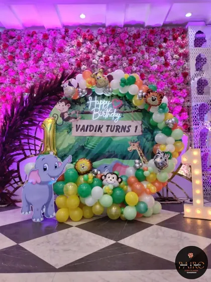 Let's get wild with a fun jungle theme. This first birthday setup for Vaidik features a circular backdrop with jungle prints, character cutouts of an elephant and other animals, a "Happy Birthday" neon sign, and a vibrant green and orange balloon arrangement.