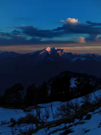The last light of the sun kisses the top of a snowy peak. This fleeting moment, known as alpenglow, paints the mountain in a soft, warm light against the darkening sky.
