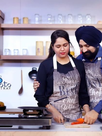A fun and relatable pre-wedding photo in a modern kitchen set. The couple wears matching aprons and pretends to cook together, showing their partnership in a casual setting.
