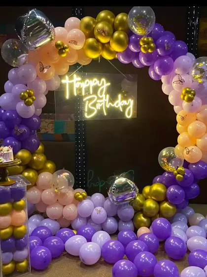 A vibrant birthday party setup with a purple, peach, and gold balloon ring. The decoration is enhanced with a neon sign and butterfly accents for a touch of whimsy.