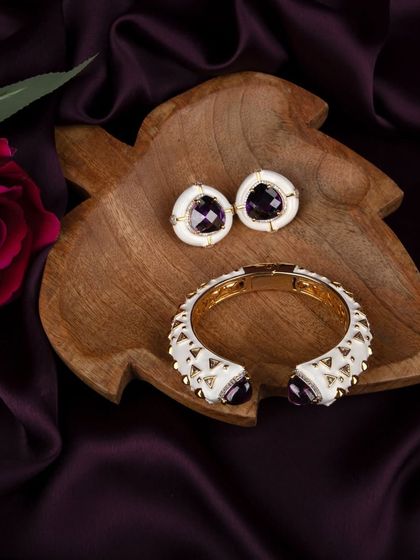 A contemporary white enamel bracelet and earring set with purple stones, styled on a leaf-shaped wooden tray with a rose for a natural, elegant look.