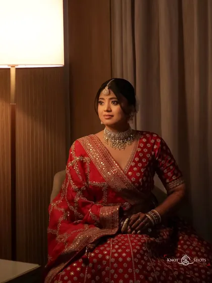 A full-length portrait of the bride seated, her red saree draped beautifully. The warm lamplight creates a cozy and elegant atmosphere.