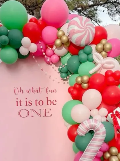 "Oh what fun it is to be ONE." A sweet, candy-themed Christmas birthday backdrop in pink, red, and green, featuring candy cane and gift box details on the balloon garland.