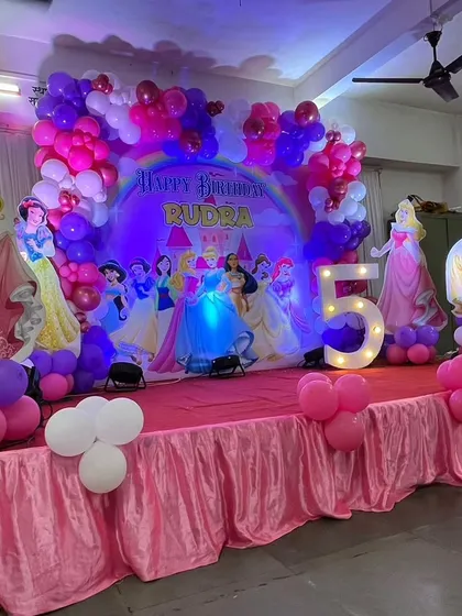 A grand Disney Princess stage for a 5th birthday party. The setup includes a large backdrop with all the princesses, a purple and pink balloon arrangement, and a light-up marquee number '5'.